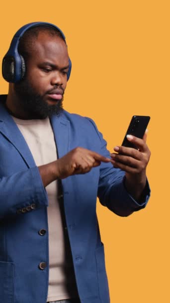 在连接智能手机的蓝牙耳机上听音乐的非洲裔美国男人的垂直视频 Bipoc人员从播放列表中选择歌曲 在录音室背景下被隔离 — 图库视频影像
