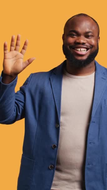 垂直ビデオ アフリカ系アメリカ人の男は陽気に挨拶手のジェスチャーを行います 幸せなBipocの人物の肖像画 誰かに挨拶するために腕を上げる ジェスチャー イエロースタジオの背景に隔離された カメラA — ストック動画