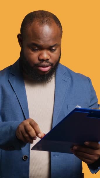 垂直视频 人浏览剪贴板上的财务审计报告 对高收入 工作室背景感到满意 快乐的非洲裔美国人在笔记本上阅读文书工作 对数字感到满意 — 图库视频影像