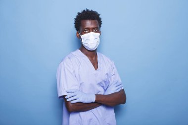Stüdyoda, kollarını kavuşturmuş, maske ve eldiven takmış hemşire kameraya bakıyor. Tıbbi asistan olarak çalışan siyahi bir adam, izole edilmiş arka planda duruyor, covid 19 salgınından korunuyor..