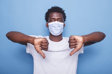 Afro-Amerikalı yetişkin iki eliyle baş parmak işareti gösteriyor. Siyah adam parmaklarıyla hareket ediyor kameraya bakıyor ve koronavirüsten korunmak için maske takıyor..