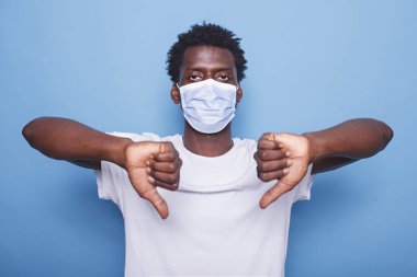 African American adult showing thumbs down sign with both hands. Black man gesturing with his fingers while looking at camera and wearing face mask to protect from coronavirus epidemic. clipart