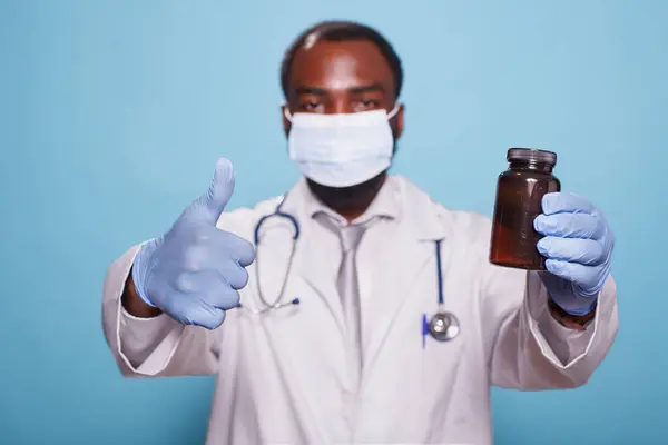 戴口罩的医学专家的画像 在抓起药瓶时露出大拇指 带听诊器和手套的黑人医生批准手部处方止痛药 图库照片