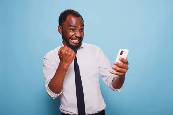 ホワイトシャツの黒人男性が携帯電話で勝利したオンラインモバイルゲームをスクリーンで緊張させた アフリカ系アメリカ人ゲーマーが初めてスマートフォンでモバイルアプリをテスト ロイヤリティフリーのストック写真