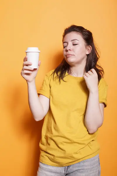 疲惫的女人穿着休闲服在孤立的橙色背景下摆姿势 拿着咖啡杯 闭着眼睛放松 年轻的黑发女士睡意朦胧的表情表现出疲倦和疲倦 图库图片