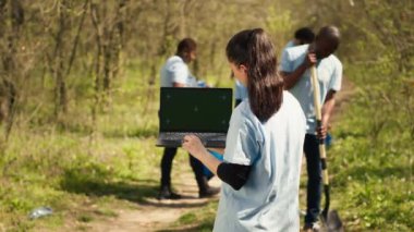 Çevre koruma gönüllüsü ekibiyle birlikte ormanı temizlemeye çalışan yeşil ekran bir dizüstü bilgisayar tutuyor. Doğa aktivistleri izole edilmiş maketler kullanır ve çöp toplarlar. Kamera A.
