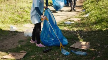 Küçük bir kız, plastik atık ve çöpleri geri dönüşümle çevreyi temizleyerek, bir pençe aleti ve çöp torbalarıyla ormanda çöp topluyor. Çocuk ekosistemi korumayı öğreniyor. Kamera A.
