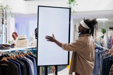 Müşteri mağazadaki kıyafetleri karıştırmak için boş beyaz ekran kullanıyor. Afro-amerikan tarzı bir adam alışveriş merkezinden kıyafet alırken interaktif boş ekrana dokunuyor.