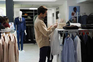Elbise mağazası müşterisi askıda resmi tişört tutuyor ve boyut etiketine bakıyor. Giyim kuşamını kontrol eden ve alışveriş merkezinden şık kıyafetler seçen genç bir adam.