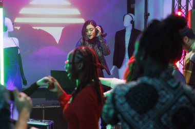 Gece kulübündeki disko partisinde sahne alan kadın DJ. Asyalı müzisyen elinde kulaklıkla elektronik müzik eşleştirirken kalabalık dans pistinde dans ediyor.