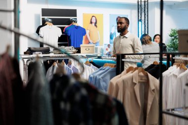 Afro-Amerikalı bir adam giyim mağazasında dikilirken giyim ve gezinme rafı satın alıyor. Alışveriş merkezi moda butik müşterisi askılarda günlük kıyafet seçiyor.