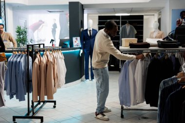 Afro-Amerikalı bir adam gömlek ölçüsünü ararken giyim mağazasında resmi kıyafet seçiyor. Alışveriş merkezinde asılı giysileri olan butik müşteri tarama rafı.
