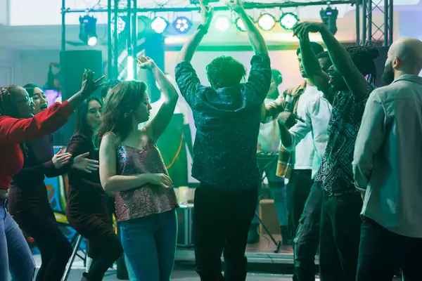 stock image Active people dancing and raising hands while partying in nightclub. Young men and women moving to electronic music beats and relaxing on crowded dancefloor at discotheque