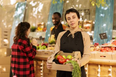 Neşeli kadın meyve ve sebze satıcısı market sahibi taze organik ürün kutusu gösteriyor ve kameraya bakıyor. Ypung çiftçisi yol kenarından sağlıklı yerel ürünler satıyor, ekolojik ürünler..