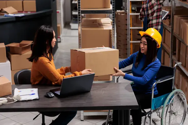 Asian woman in a wheelchair managing postal service orders and coworking with logistics manager. Storehouse employees handling package distribution in inclusive workplace