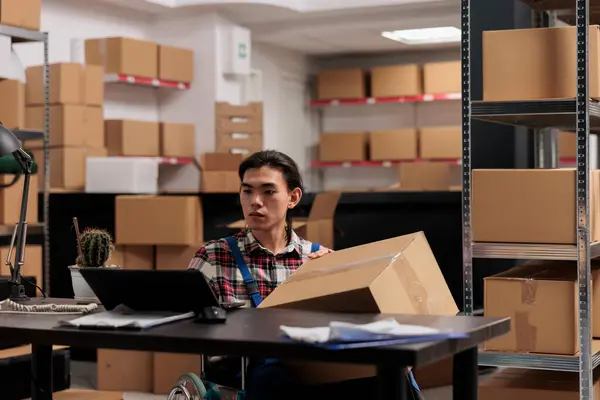 Warehouse Employee Wheelchair Processing Order Laptop While Holding Cardboard Box Stockafbeelding