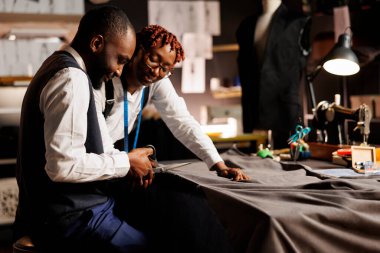 Experienced african american atelier shop coworkers meticulously cutting refined textile material for customer comissioned suit. Expert suitmakers manufacturing bespoke sartorial outfit clipart