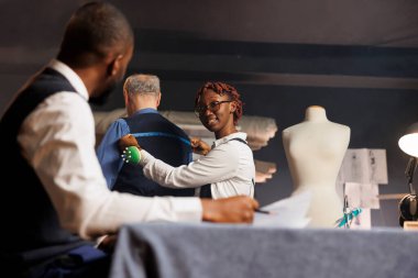 Senior businessman getting back measurements taken by meticulous needleworker in atelier workspace. Precise skilled tailor sizing up elderly client proportions for handmade bespoken suit clipart