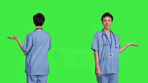 医療アシスタントは 何かを脇に置いてウェブコマーシャルを行い 緑色の背景に対して広告を作成します スクラブを持つ若い看護師は 左側または右側に何かを示しています カメラ — ストック写真