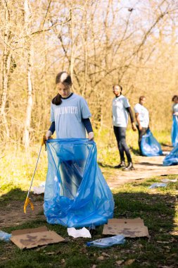 Kamu hizmeti gönüllüleri atıkları azaltır ve orman ortamını temizler, çöpleri mavi çöp torbalarında toplar. Genç çocuk plastik atıkları atarak çevrenin korunmasına yardım ediyor.