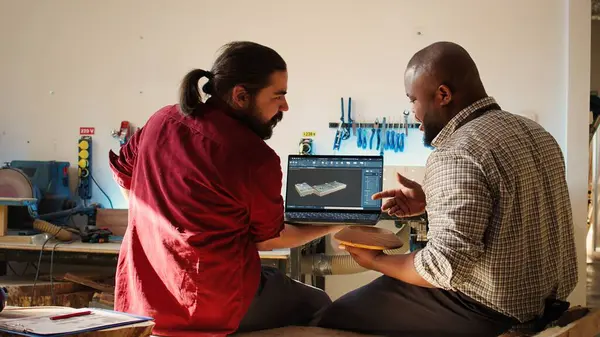 stock image African american manufacturer and colleague comparing wood art piece with schematic displayed on CAD software on laptop. Woodworking professionals crosschecking wooden object with blueprint, camera B