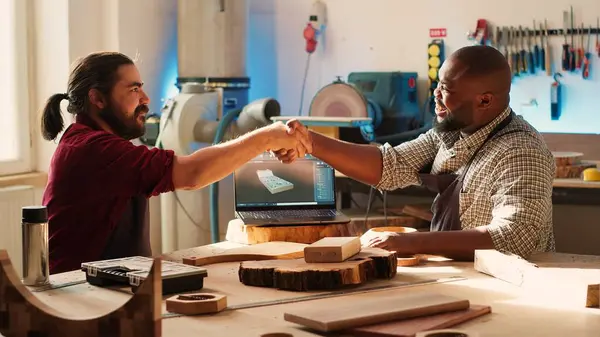 stock image Cabinetmaker shaking colleague hand after finishing using CAD software on laptop to design wooden objects. Carpenters congratulating each other for job well done in furniture assembling shop, camera A