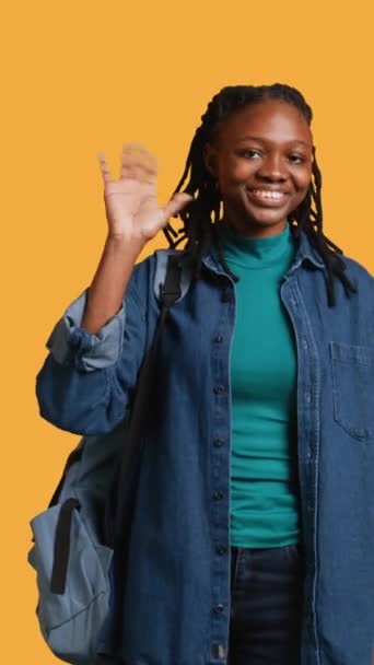 バックパックを保持するチッパーガールの垂直ビデオ肖像画 手を振り ジェスチャー イエロースタジオの背景の上に隔離 ジョリー アフリカ系アメリカ人学生と学校のリュックサックの挨拶手 — ストック動画