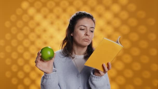 女人读书 享受新鲜的苹果 健康地生活 独立于工作室背景之外 书虫以新奇的绿果为乐 享受生物小吃 相机B — 图库视频影像