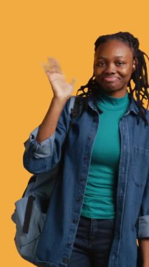 Mutlu bir kadının el sallayan, el sallayan, el sallayan, el kol hareketi yapan, sarı stüdyo arka planında izole edilmiş dikey video görüntüsü. Sırt çantalı neşeli BIPOC genci selamlama hareketi yapıyor, kamera B