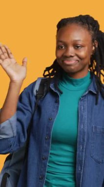 Mutlu bir kadının el sallayan, el sallayan, el sallayan, el kol hareketi yapan, sarı stüdyo arka planında izole edilmiş dikey video görüntüsü. Sırt çantalı neşeli BIPOC genci selamlama hareketi yapıyor, kamera A