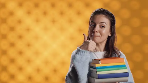 stock image Portrait of happy woman with stack of books in hands showing thumbs up, studio background. Joyous bookworm holding pile of novels, feeling upbeat, doing positive hand gesturing, camera A