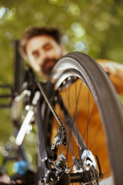 Detailed shot of bicycle wheel rubber chain being examined and maintained by young healthy caucasian man outdoor. Image showing close-up view of bike tire and pulley wheels in yard. clipart