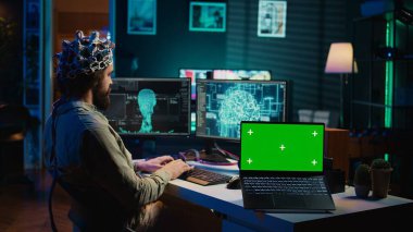 Engineer with EEG headset on programming brain transfer into computer virtual world next to green screen laptop. Transhumanist using neuroscience to gain digital soul, mockup device, camera B clipart