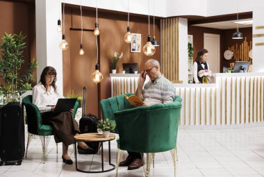 Emekli yaşlı kadın otel lobisindeki bir kanepede oturuyor ve bilgisayarını kullanıyor. Yaşlı bayan kablosuz bilgisayarını karıştırırken üst düzey bir erkek müşteri dinlenme odasında kitap okuyor..