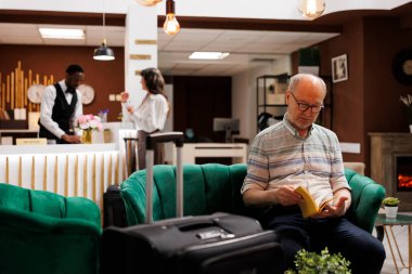 Speaking to young concierge at main desk of luxury resort is caucasian female traveller. While old woman completes booking process at counter an elderly man reading a book relaxes in hotel lobby. clipart
