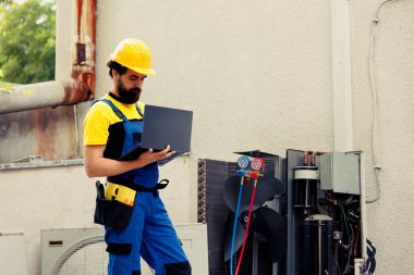 Hasarlı elektrik parçalarını aramak için harici Hvac sistem rutin bakımı için eğitimli tamirci görevlendirildi. Yoğunlaştırıcı güvenlik kontrolü yapan titiz elektrikçi, dizüstü bilgisayara rapor yazıyor.