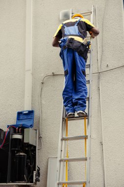 Profesyonel üniformalı uzman tamirci çatıdaki havalandırma sisteminin bakımını yapmak için teknik ekipman katlama merdivenine tırmanıyor. Klimayı kontrol etmek için uzman bir tamirci görevlendirildi.