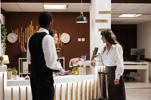 stock image Retired senior woman at front desk doing check-in process in exclusive hotel lobby. While bellboy helps with suitcase in lounge area, concierge assists elderly female tourist with room reservation.