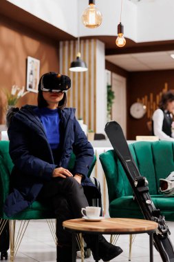 In hotel reception, young female traveler adorned in snow gear including winter jacket, attentively using vr glasses. Asian woman having vr headset sits in lounge area of ski mountain resort. clipart