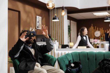 Male tourist wearing winter jacket is using a vr headset in lounge area of ski mountain resort. While waiting in hotel lobby man with beard utilizes vr goggles for research on wintersports activity. clipart