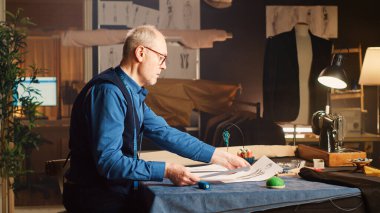 Deneyimli bir terzi dikiş ekipmanı kullanarak atölyedeki kıyafet tasarımları üzerinde çalışıyor. Kıdemli tasarımcı el yapımı tasarımlar için çizimler yapıyor. Üçayak atışı.