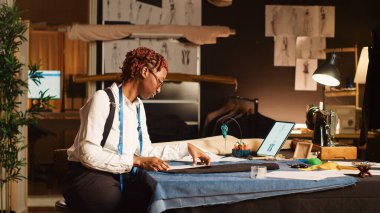 Kadın terzi, tablet ve kağıtlardaki çizimleri kullanarak giysiler tasarlıyor, modern tasarım ile yeni moda koleksiyonu üzerinde çalışıyor. Genç modacı terzilik atölyesinde elbise dizayn ediyor. Üçayak atışı.