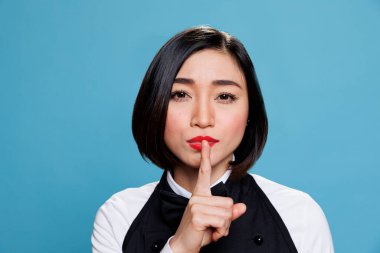 Hotel young asian woman receptionist making silent gesture, asking to keep secret portrait. Restaurant waitress wearing uniform holding forefinger on lips and looking at camera clipart