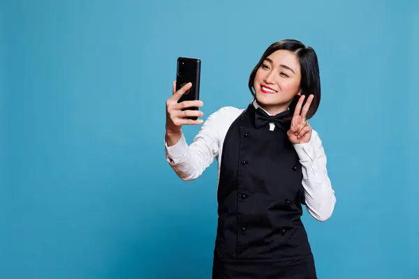 Smiling Asian Waitress Holding Smartphone Looking Front Camera Show Peace Stok Lukisan  