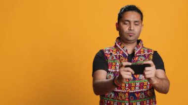 Akıllı telefondaki video oyunlarıyla eğlenen mutlu Hintli adam boş vakitlerin tadını çıkarıyor. Oyuncu telefonda oyundan zevk alıyor, düşmanları yenerken eğleniyor, stüdyo arka planında izole edilmiş, kamera A