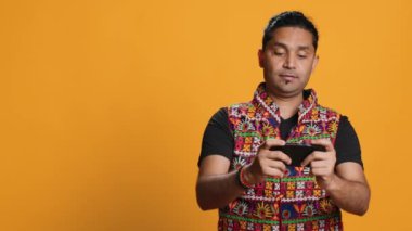 Geleneksel giyimli neşeli Hintli insan akıllı telefonuyla video oyunlarıyla eğleniyor. Neşeli oyuncu telefondan keyif alıyor, çevrimiçi eğleniyor, stüdyo arkaplanı, kamera A