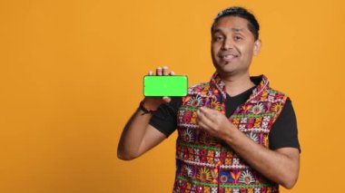 İçerik yaratıcısı yeşil ekran cep telefonunu sunuyor, stüdyo arka planında izole edilmiş. Hintli kişi internet kanalı için kopyalama alanı modelleme akıllı telefon aygıtı, kamera A 'yı inceliyor