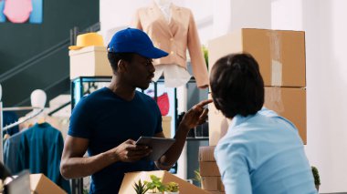 Mağaza çalışanı nakliye raporunu imzalıyor, alışveriş merkezinde Afrikalı Amerikan kuryesiyle lojistiği tartışıyor. Modern butikte teslimat için müşteri paketleri hazırlayan şık bir yönetici