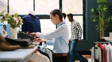 Asyalı işçi şık bir kravat takıyor, modern butikte aksesuarları rafa koyuyor. Perakende satış elemanı mağazayı açılışa hazırlıyor, alışveriş merkezinde şık kıyafetlerle çalışıyor. Moda konsepti