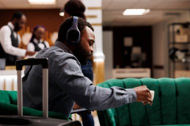 Ciddi bir Afro-Amerikan işadamı, otel odasının hazır olmasını beklerken kahve içerken lobide bavuluyla oturuyor. İnsanlar ve seyahat, iş gezisi konsepti